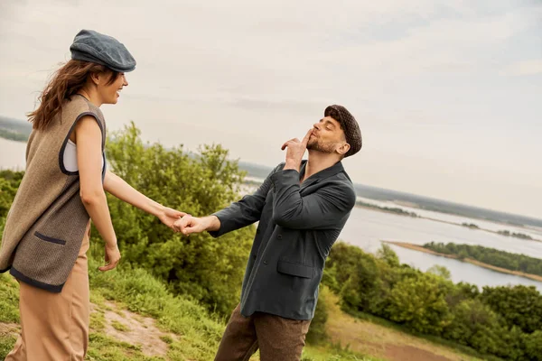 Fashionable man in newsboy cap and jacket showing secret gesture and holding hand of cheerful girlfriend while standing with nature at background, fashion-forwards in countryside — Stock Photo