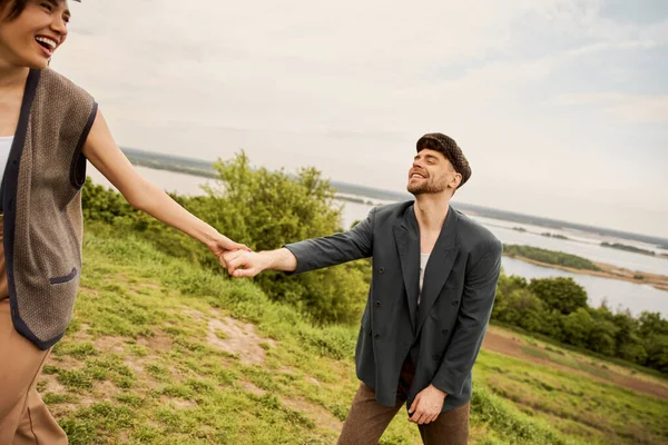 Cheerful and stylish woman in vest holding hand of bearded boyfriend in newsboy cap and jeans with scenic nature and overcast sky at background, fashion-forwards in countryside — Stock Photo