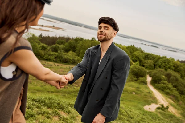 Smiling and bearded man in stylish newsboy cap and jacket holding hand of blurred brunette girlfriend while standing with blurred nature and sty at background, fashion-forwards in countryside — Stock Photo