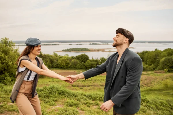 Positive and fashionable brunette woman in newsboy cap and vest holding hand of bearded boyfriend in jacket while standing with scenic landscape at background, fashionable couple in countryside — Stock Photo