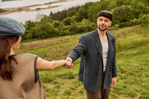 Smiling bearded man in newsboy cap and jacket holding hand of brunette girlfriend in vest while standing with nature at background, fashionable couple in countryside — Stock Photo