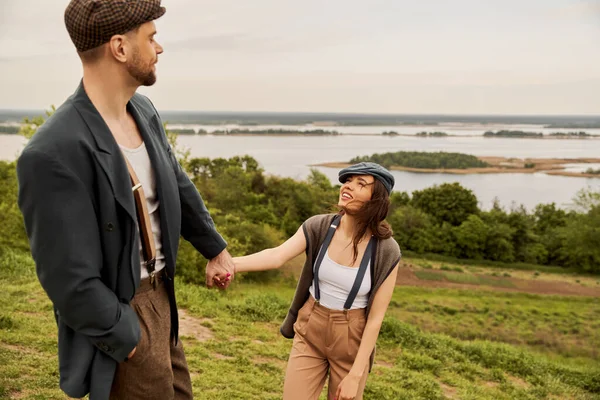 Cheerful and fashionable brunette woman in newsboy cap and suspenders holding hand of bearded boyfriend in jacket and standing with scenic landscape at background, fashionable couple in countryside — Stock Photo