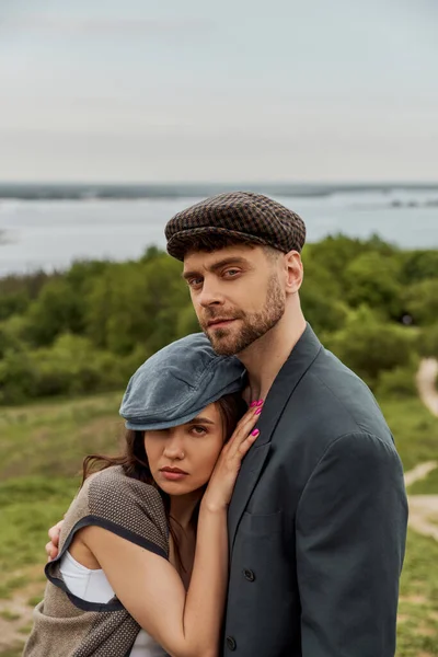 Portrait of confident bearded man in jacket hugging brunette girlfriend in newsboy cap and vest and looking at camera while standing with landscape at background, fashionable couple in countryside — Stock Photo