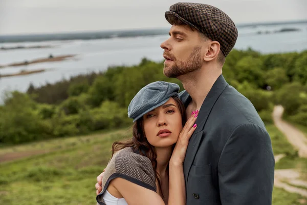 Brunette et femme à la mode en bonnet de newsboy et gilet étreignant petit ami barbu en veste et debout avec paysage pittoresque en arrière-plan, couple à la mode dans la campagne — Photo de stock