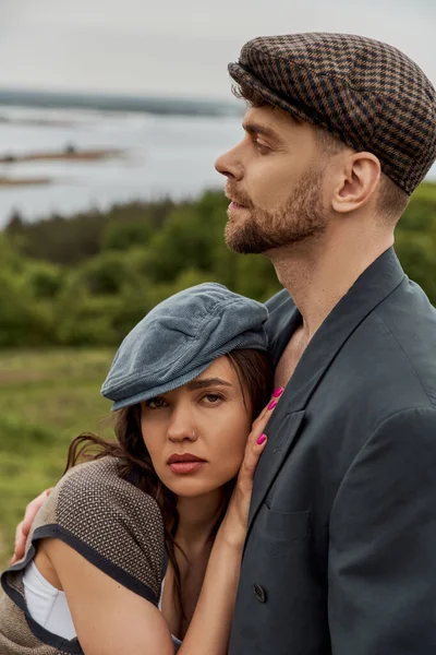 Portrait of trendy brunette woman in vest and vintage outfit hugging bearded boyfriend in jacket and newsboy cap with rural landscape at background, fashionable couple in countryside — Stock Photo