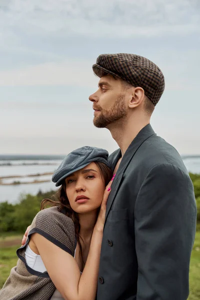 Trendy and brunette woman in newsboy cap and vest hugging bearded boyfriend in jacket and looking at camera with rural landscape at background, fashionable couple in countryside — Stock Photo