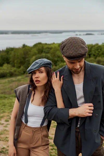 Fashionable brunette woman in suspenders and newsboy cap touching bearded boyfriend in jacket and looking at camera with blurred nature at background, stylish pair amidst nature — Stock Photo