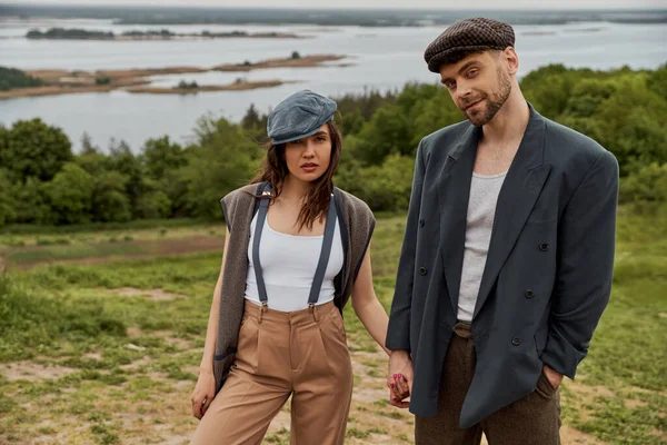 Confident and bearded man in jacket holding hand of fashionable girlfriend in newsboy cap and suspenders and looking at camera while standing with nature at background, stylish pair amidst nature — Stock Photo