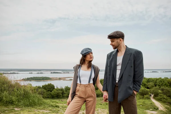 Fashionable and bearded man in newsboy cap and jacket holding hand of brunette girlfriend in suspenders and vest and standing together with nature at background, stylish pair amidst nature — Stock Photo