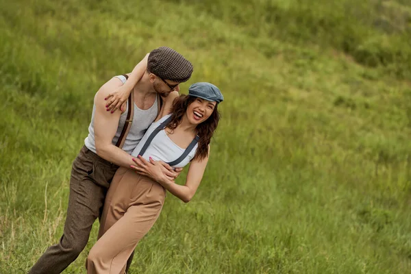 Fashionable man in vintage outfit and sunglasses hugging cheerful girlfriend in suspenders and newsboy cap while spending time together and standing on grassy lawn, stylish pair amidst nature — Stock Photo
