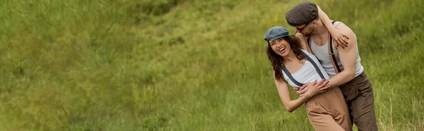 Homme barbu à la mode en tenue vintage et lunettes de soleil étreignant petite amie positive dans un bonnet de newsboy et debout sur une pelouse gazonnée floue à l'arrière-plan, paire élégante au milieu de la nature, bannière — Photo de stock