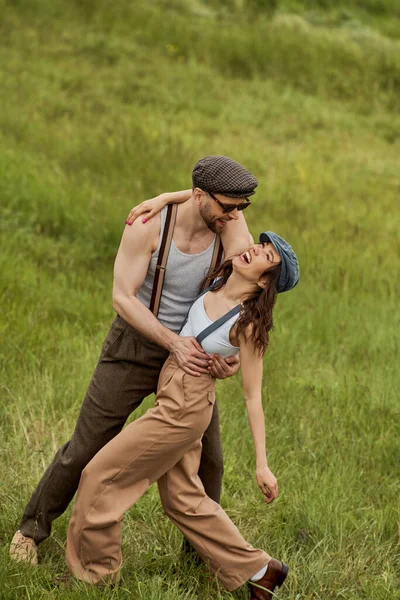 Cheerful bearded man in sunglasses and vintage outfit hugging girlfriend in newsboy cap and suspenders while standing together on grassy lawn at background, stylish pair amidst nature — Stock Photo