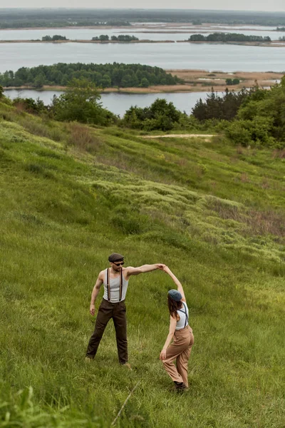 High angle view of fashionable man in sunglasses and newsboy cap having fun with brunette girlfriend in vintage outfit and standing on grassy hill at background, stylish couple enjoying country life — Stock Photo