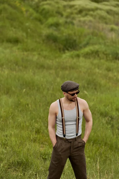 Trendy man in vintage-inspired outfit sunglasses and suspenders holding hands in pockets of pants while standing on blurred grassy meadow at background, man enjoying country life — Stock Photo
