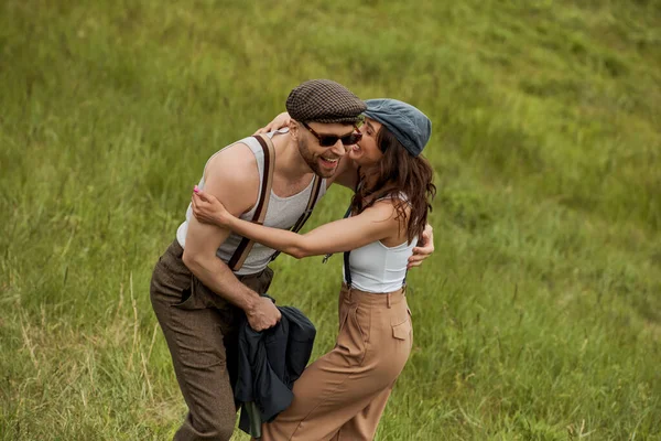 Cheerful brunette woman in vintage outfit and newsboy cap hugging and having fun with bearded boyfriend in sunglasses and standing on grassy hill, stylish couple enjoying country life — Stock Photo