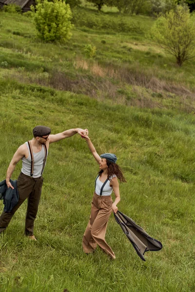 Side view of cheerful brunette woman in vintage clothes holding vest and having fun with boyfriend in newsboy cap and sunglasses on grassy meadow, stylish couple enjoying country life — Stock Photo