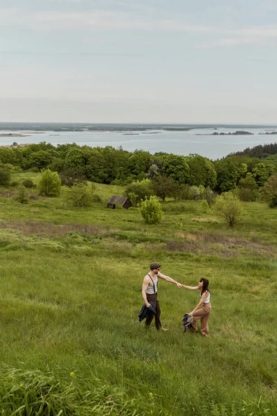 Вид позитивной и стильной пары в винтажных нарядах, держащейся за руки и стоящей на холме с сельским пейзажем на заднем плане, стильная пара, наслаждающаяся деревенской жизнью — стоковое фото