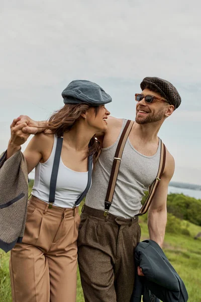 Smiling bearded man in vintage outfit and newsboy cap hugging brunette girlfriend with suspenders and walking with scenic landscape at background, stylish couple enjoying country life — Stock Photo