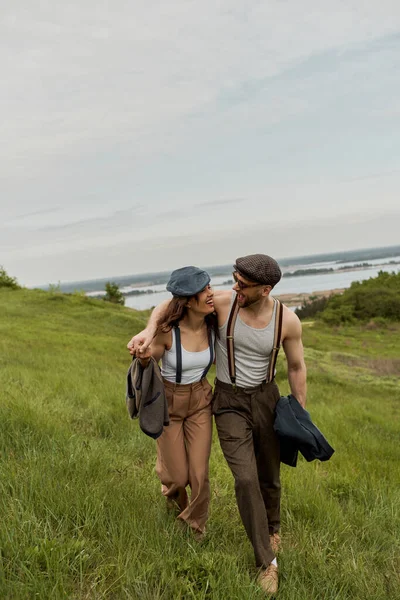 Cheerful and fashionable romantic couple in newsboy caps, vintage outfits and suspenders hugging and walking together on grassy hill at background, trendy couple in the rustic outdoors — Stock Photo