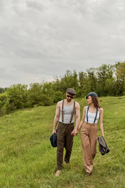 Fashionable brunette woman in newsboy cap and suspenders holding hand of bearded boyfriend in sunglasses and talking while walking together on grassy lawn, trendy couple in the rustic outdoors — Stock Photo