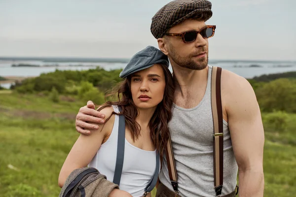 Stylish man in newsboy cap and sunglasses hugging brunette girlfriend in suspenders holding vest and looking at camera while standing with field at background, trendy couple in the rustic outdoors — Stock Photo