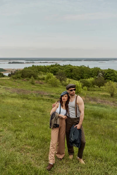 Homem na moda em óculos de sol e boné de jornal abraçando namorada morena e segurando jaqueta enquanto passa o tempo e de pé no campo gramado, casal na moda no rústico ao ar livre — Fotografia de Stock