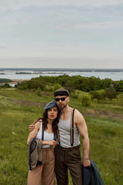 Модна романтична пара в шапочках і підвісках, що тримають піджак і жилет і обіймаються, стоячи на розмитому трав'янистому полі на фоні, модна пара на сільському відкритому повітрі — стокове фото