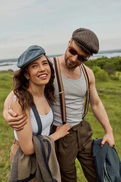 Homem elegante em óculos de sol e suspensórios abraçando namorada morena alegre e jaqueta segurando enquanto caminha no campo gramado turvo, casal na moda no rústico ao ar livre — Fotografia de Stock