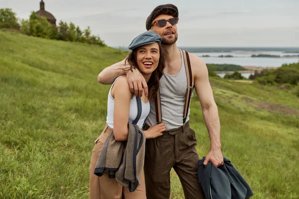 Homem alegre e barbudo em óculos de sol e roupa vintage abraçando namorada morena em boné de jornal e de pé juntos na paisagem rural, casal na moda no rústico ao ar livre — Fotografia de Stock