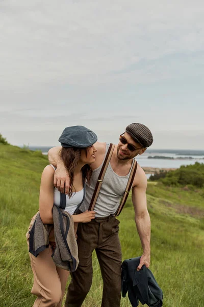 Lächelnder und trendiger Mann mit Sonnenbrille umarmt brünette Freundin in Nachrichtenmütze und Hosenträgern und verbringt Zeit auf verschwommenem Rasen mit bewölktem Himmel im Hintergrund, stilvolle Partner in ländlicher Flucht — Stockfoto