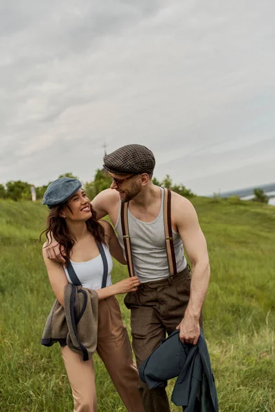 Fashionable bearded man in sunglasses and newsboy cap hugging cheerful brunette girlfriend and holding jacket while walking together in rural landscape, stylish partners in rural escape — Stock Photo