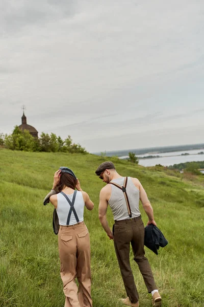 Fashionable romantic couple in vintage outfits, newsboy caps and suspenders walking together on grassy hill with cloudy sky at background, stylish partners in rural escape, romantic getaway — Stock Photo