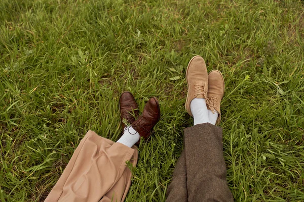 Top view of legs of romantic couple in vintage shoes and pants sitting next to each other on grassy lawn, stylish partners in rural escape, romantic getaway — Stock Photo