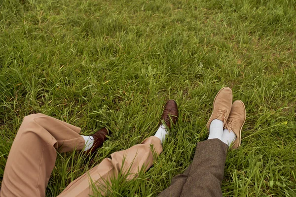 Vista superior das pernas do casal romântico em calças e sapatos retro-estilo sentados ao lado uns dos outros no prado gramado, parceiros elegantes na fuga rural, escapadela romântica — Fotografia de Stock