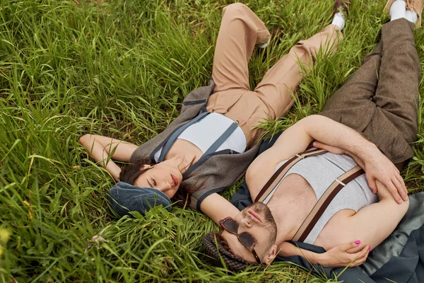 Top view of stylish brunette woman in vintage outfit and newsboy cap hugging bearded boyfriend in sunglasses while lying together and spending time on grassy meadow, stylish partners in rural escape — Stock Photo