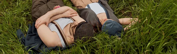 Blick aus der Vogelperspektive auf modische romantische Paare in Nachrichtenmützen und Hosenträgern, die zusammen auf einem grünen Gras liegen und entspannen, stilvolle Partner in ländlicher Flucht, Banner, romantisches Wochenende — Stockfoto