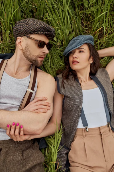 Top view of fashionable romantic couple in newsboy caps, suspenders and vintage outfits looking at each other while lying and relaxing on grassy field, stylish partners in rural escape — Stock Photo