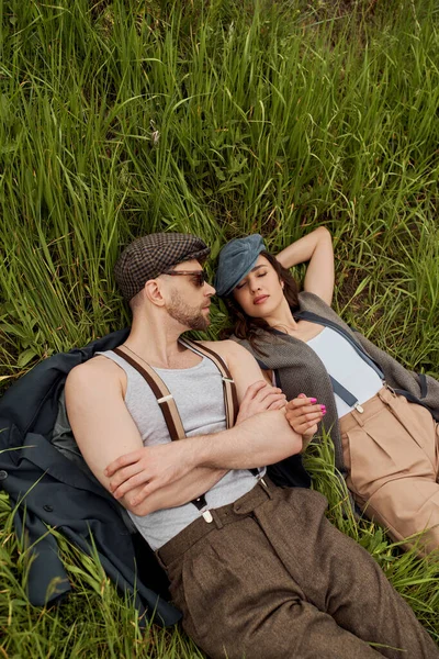 Top view of bearded man in sunglasses and susenders lying on jacket near stylish girlfriend in vintage outfit while relaxing on grassy field, fashionable couple surrounded by nature — стоковое фото