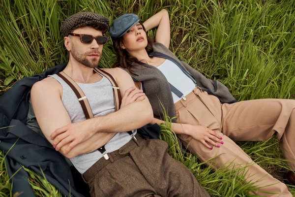 Вид зверху на модну романтичну пару в сонцезахисних окулярах, шапочках новин і вінтажний одяг, що дивиться на камеру, лежачи на трав'янистому полі, модна пара в оточенні природи — стокове фото