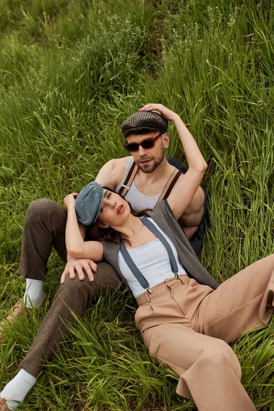 Vista de ángulo alto de la pareja romántica de moda en trajes vintage, gorras de reportero y tirantes mirando a la cámara mientras se sienta y se relaja en el campo verde, pareja de moda rodeada de naturaleza - foto de stock