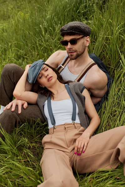 Модный мужчина в кепке газетчика и солнцезащитных очках, сидящий рядом с брюнеткой в подтяжках и винтажном наряде на зеленой траве и лугу, модная пара в окружении природы, романтический отдых — стоковое фото