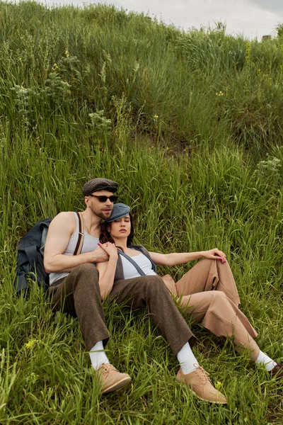Pareja romántica de moda en gorras de reportero, tirantes y trajes vintage sentados juntos en la colina con hierba verde y pasar tiempo en verano, pareja de moda rodeada de naturaleza - foto de stock