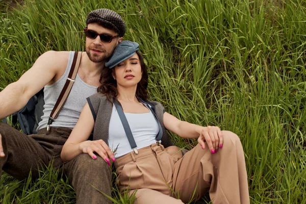 Mujer morena de moda en gorra de reportero y traje vintage cerrando los ojos mientras está sentado cerca de novio barbudo en gafas de sol en el campo de hierba en verano, pareja de moda rodeada de naturaleza - foto de stock