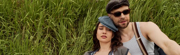 Stylish brunette woman in newsboy cap sitting with closed eyes next to bearded boyfriend in sunglasses and vintage outfit on grassy hills, fashionable couple surrounded by nature, banner — Stock Photo
