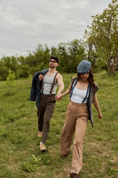 Mujer morena de moda en traje vintage y gorra de reportero cogida de la mano del novio barbudo en gafas de sol con chaqueta y caminando en el campo de hierba, pareja de moda rodeada de naturaleza - foto de stock