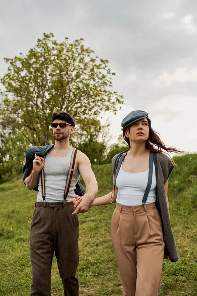 Stylish romantic couple in newsboy caps, suspenders and vintage outfits holding hands while standing with cloudy sky and green field at background, trendy twosome in rustic setting, romantic getaway — Stock Photo