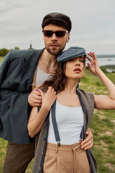 Trendy bearded man in jacket and sunglasses hugging brunette girlfriend in newsboy cap and suspenders while standing with rural landscape at background, trendy twosome in rustic setting — Stock Photo