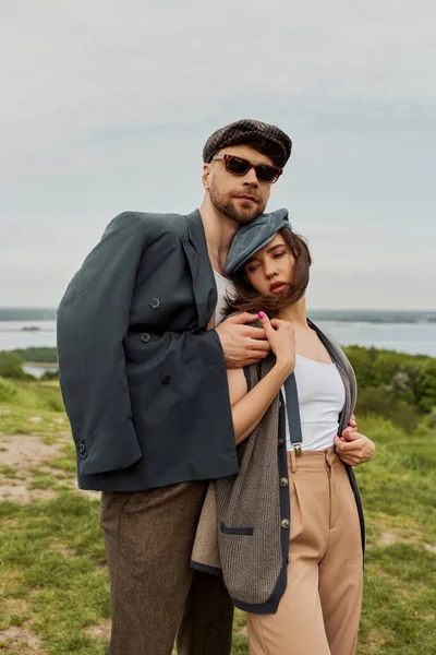 Fashionable bearded man in sunglasses and jacket hugging brunette girlfriend in newsboy cap and suspenders while spending time in rural setting, trendy twosome in rustic setting, romantic getaway — Stock Photo