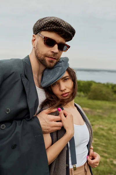 Портрет модного мужчины в солнцезащитных очках и куртке обнимающей брюнетку подружки в кепке газетчика и винтажной одежде и смотрящего в камеру с размытым пейзажем на заднем плане, романтического отдыха — стоковое фото