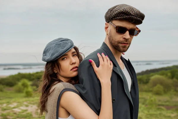 Portrait of trendy brunette woman in newsboy cap and vest hugging boyfriend in sunglasses and jacket while looking at camera with landscape at background, trendy twosome in rustic setting — Stock Photo